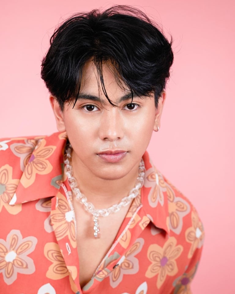 Person with black hair and bangs wearing a coral floral shirt and crystal chain necklace against a pink background