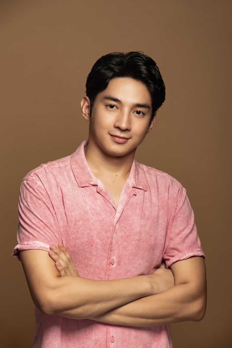 Young man with black hair wearing a pink button-up shirt, arms crossed, smiling at camera against a tan background.