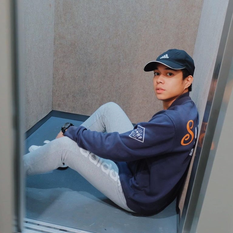 Person in navy Adidas jacket and cap sitting in a blue bathtub, looking at camera