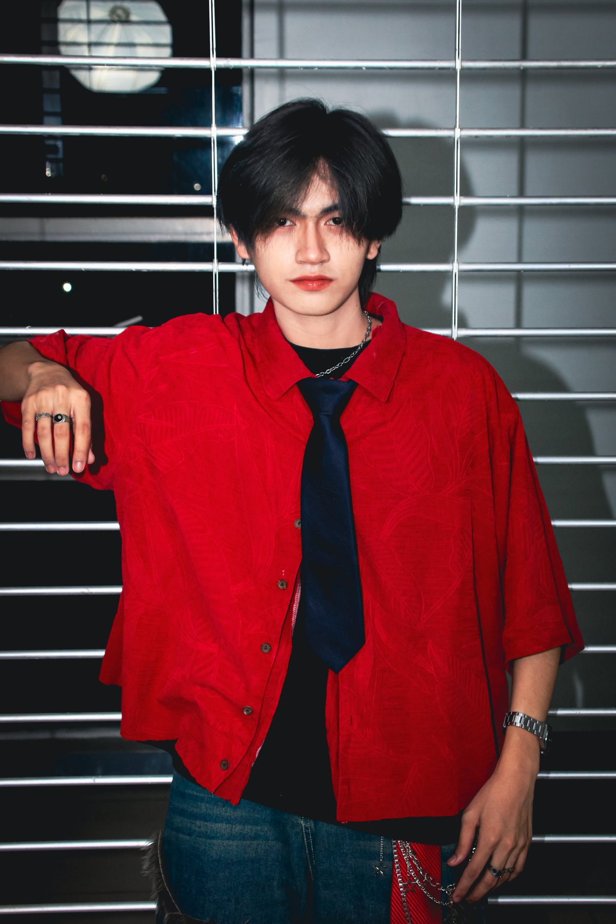 Young man with dark hair wearing a red blazer, black tie, and jeans, posing against a striped background