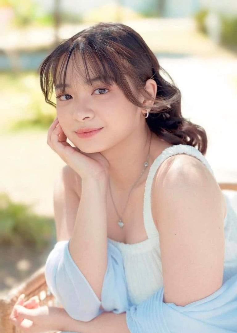 Young woman with dark hair and bangs wearing a light blue sleeveless top, posing with hand near face outdoors
