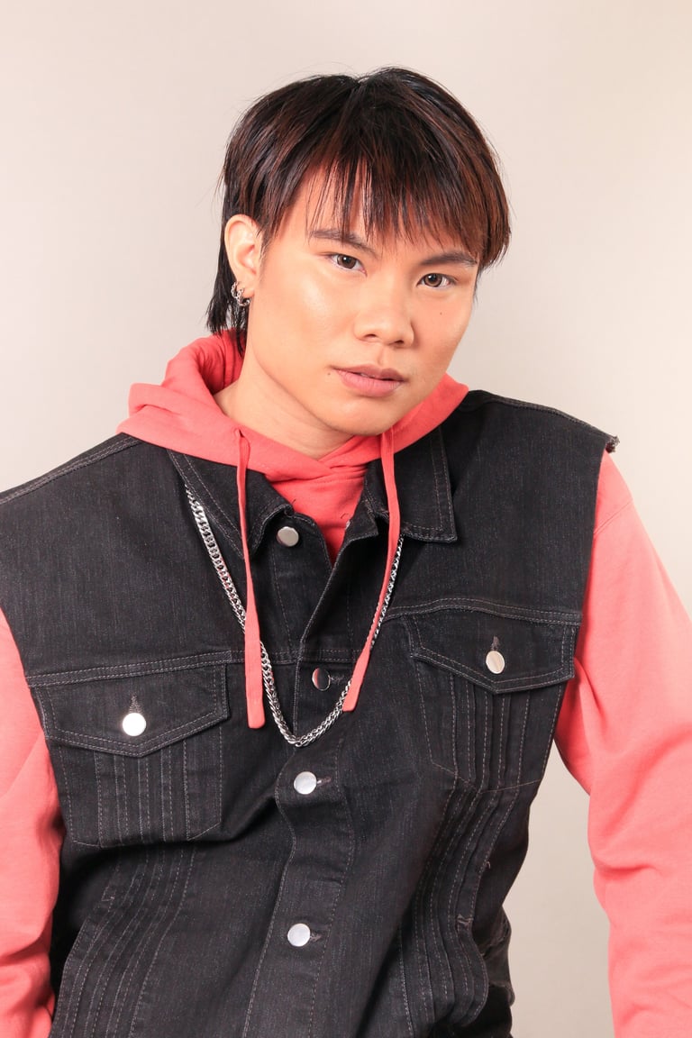 Young person with dark hair and bangs wearing a black denim vest over a pink hoodie with a chain necklace, against a light background