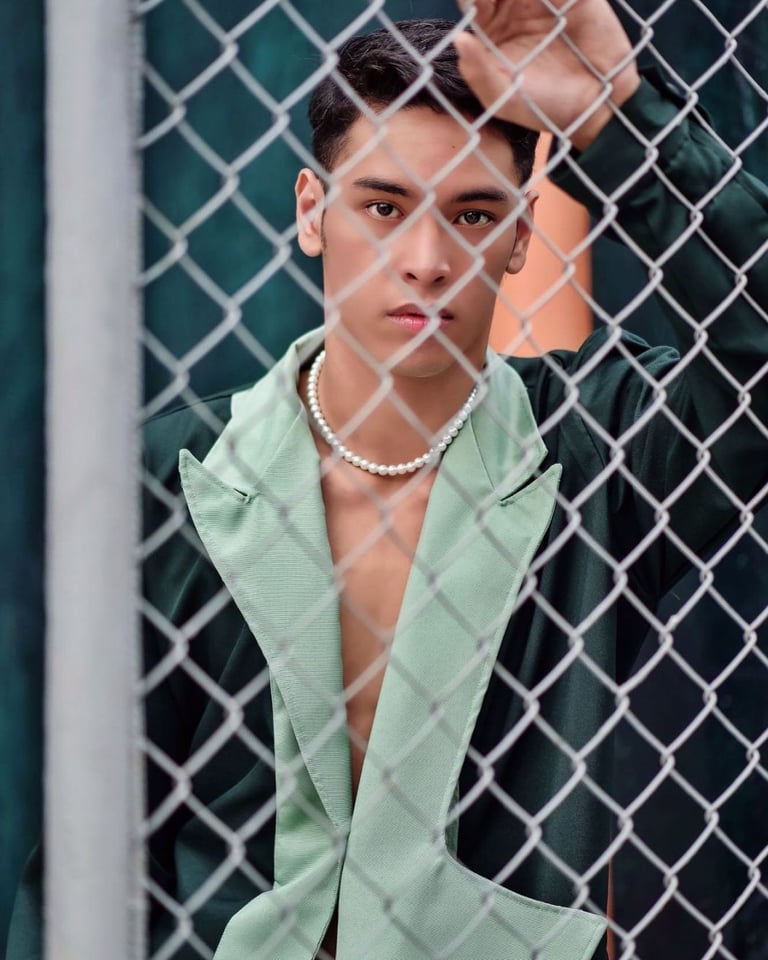 Young person wearing a light green shirt and pearl necklace posing against a chain-link fence with teal background