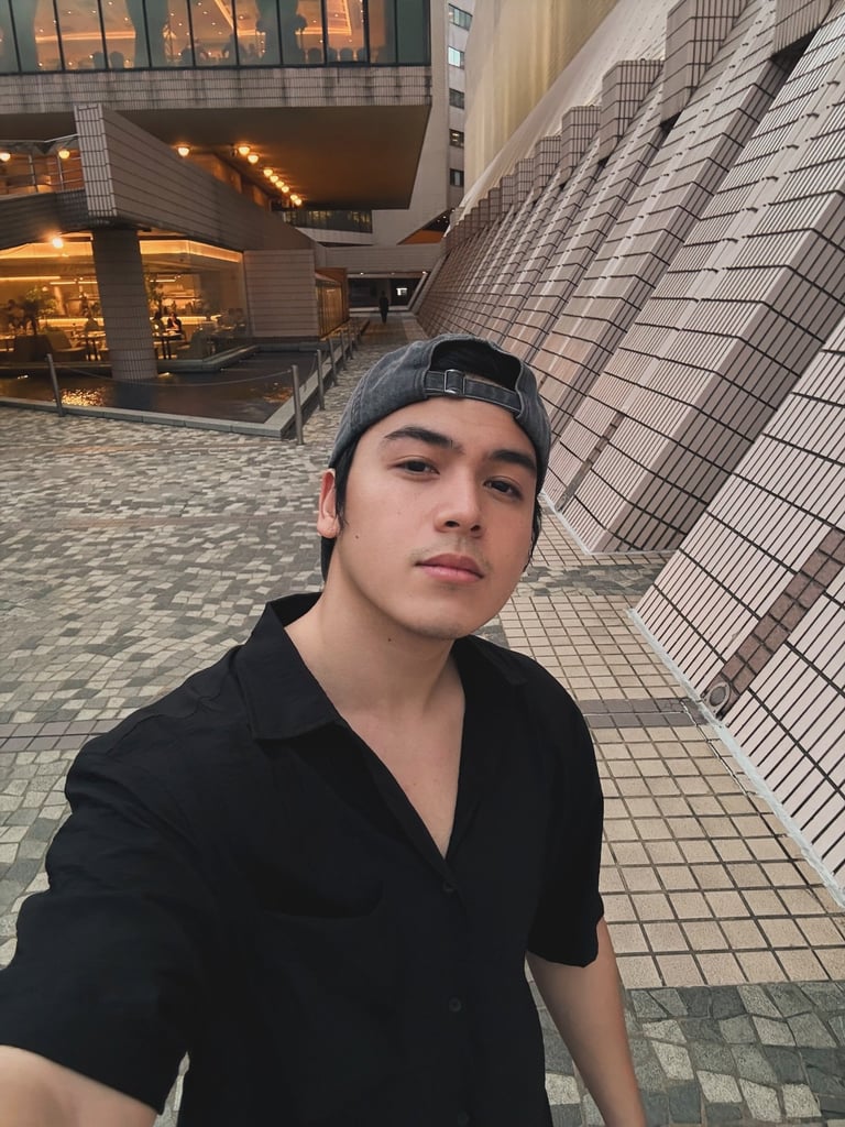 Young man wearing black shirt and cap standing in modern urban plaza with tiled buildings at dusk