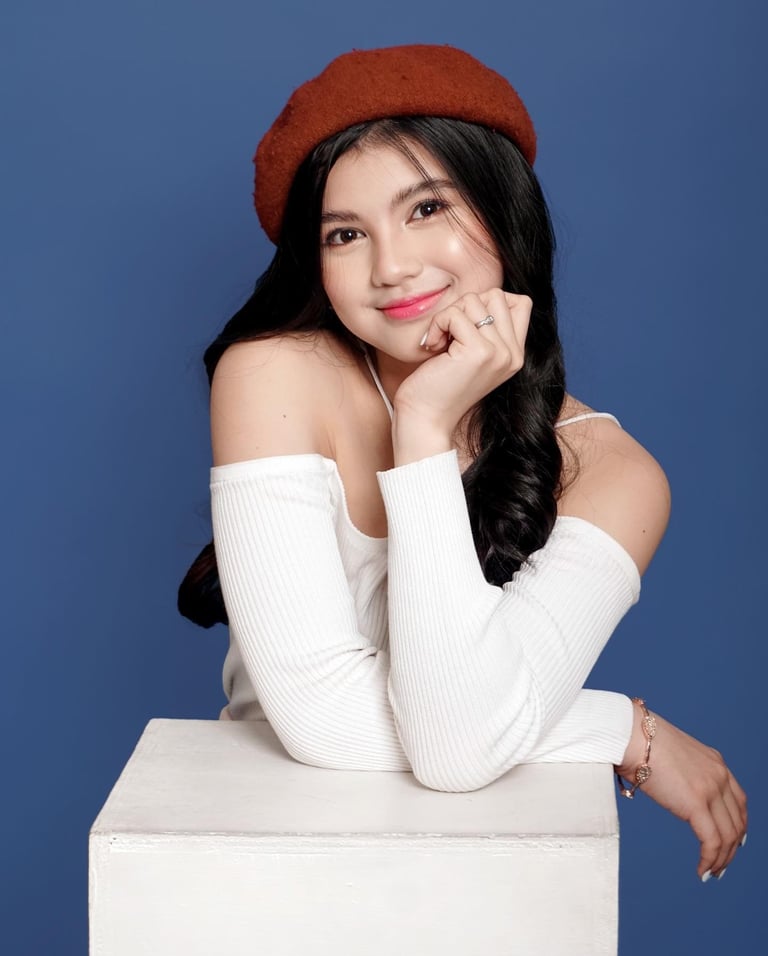 Young woman with long black hair wearing a red beret and white off-shoulder top, posing on a white cube against a blue background