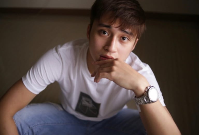 Young man in white t-shirt and jeans sitting with thoughtful expression, hand near face, wearing watch