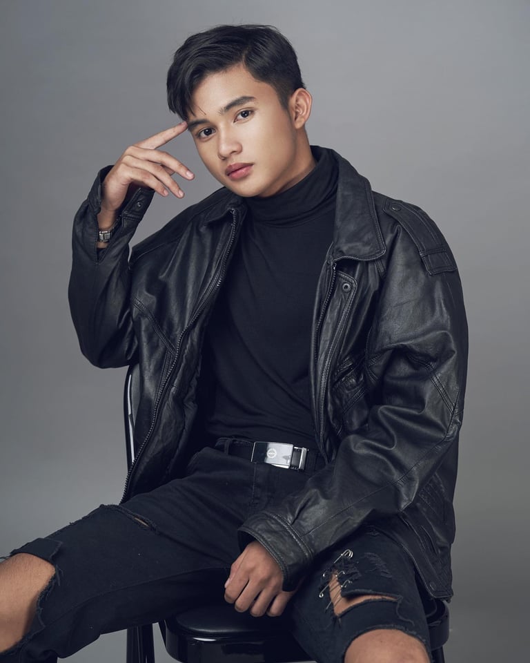 Young man wearing black leather jacket and distressed black jeans, posing against gray background with hand near face