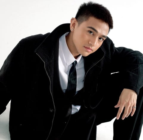 Young man in black velvet blazer with white shirt and dark tie, posed against white background looking at camera