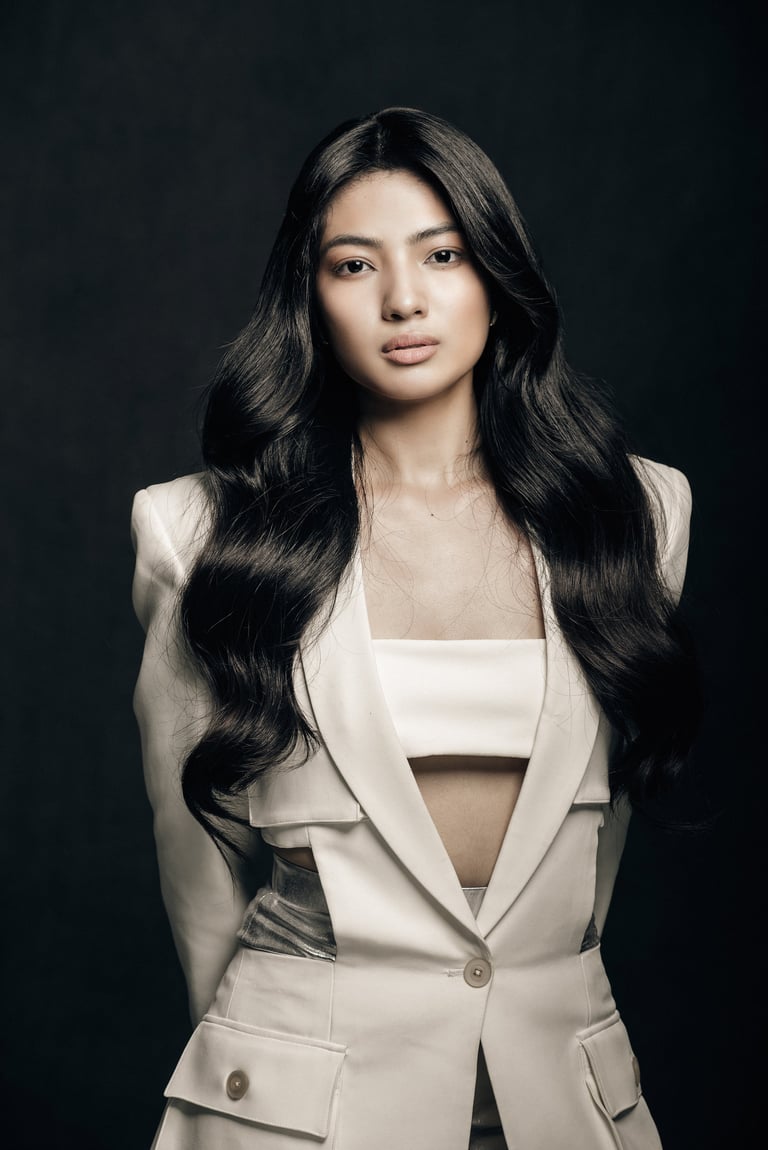 Professional portrait of a woman with long black wavy hair wearing a cream-colored sleeveless blazer against a dark background