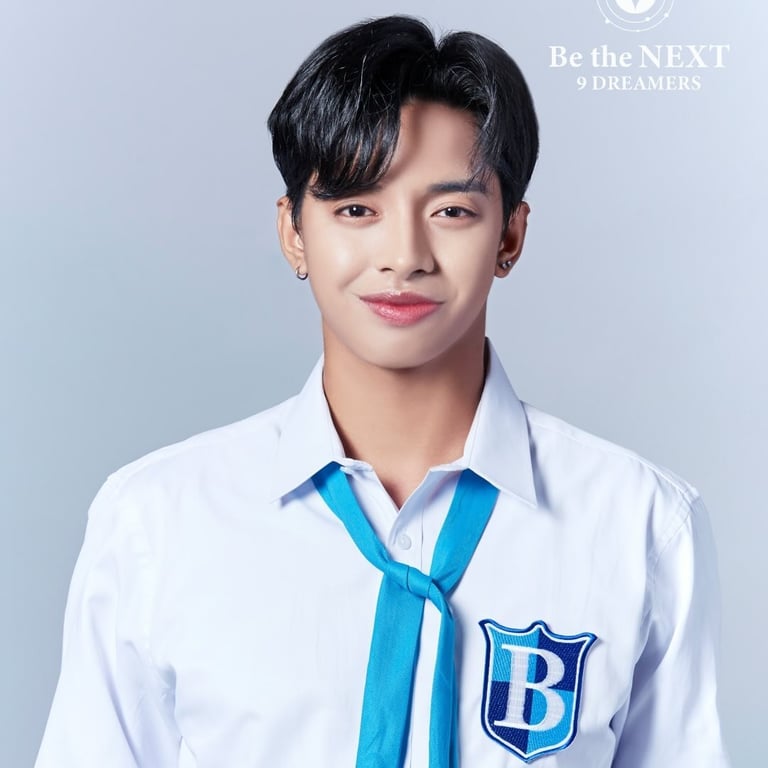 Young man in white school uniform with blue tie and B-emblazoned badge, smiling at camera against light blue background