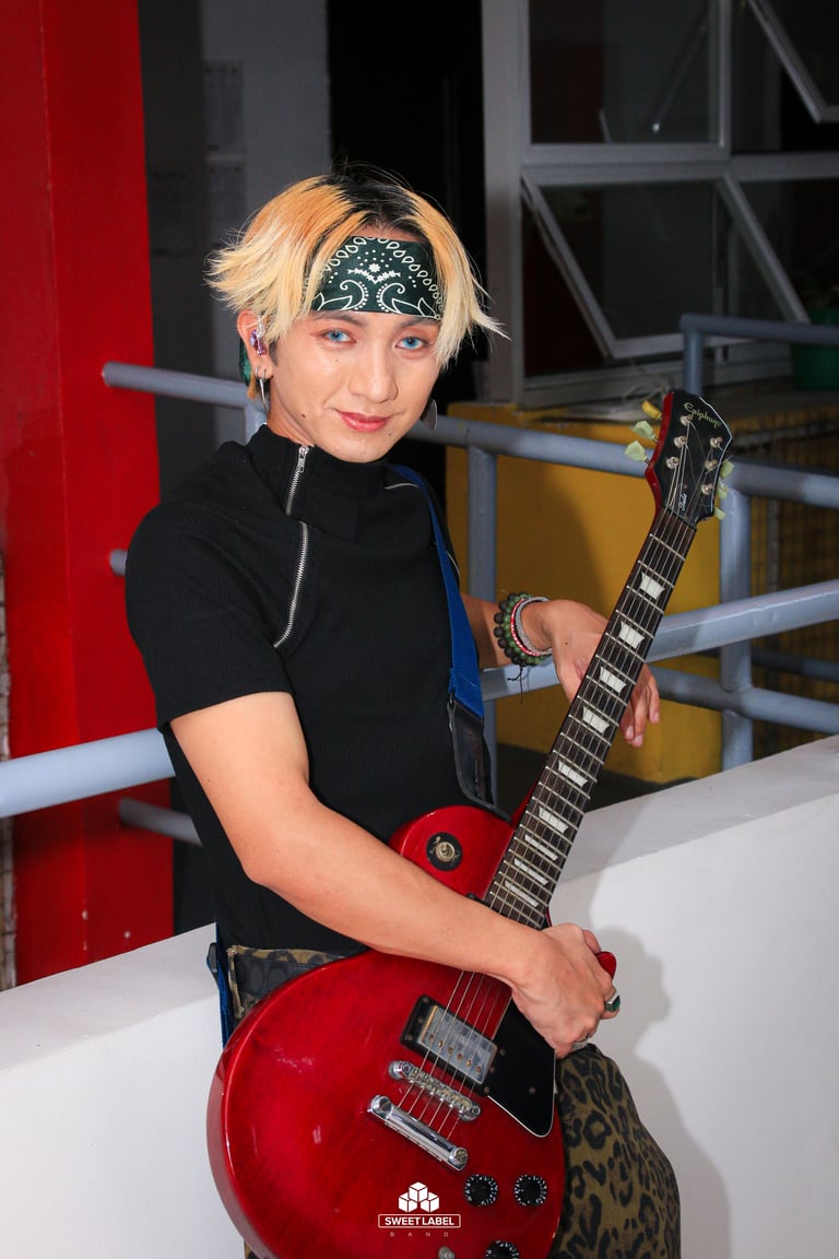 Person with blonde and black hair wearing a bandana and black shirt, holding a red electric guitar