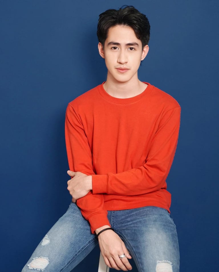 Young man wearing a red long-sleeve shirt and distressed blue jeans, sitting against a blue background
