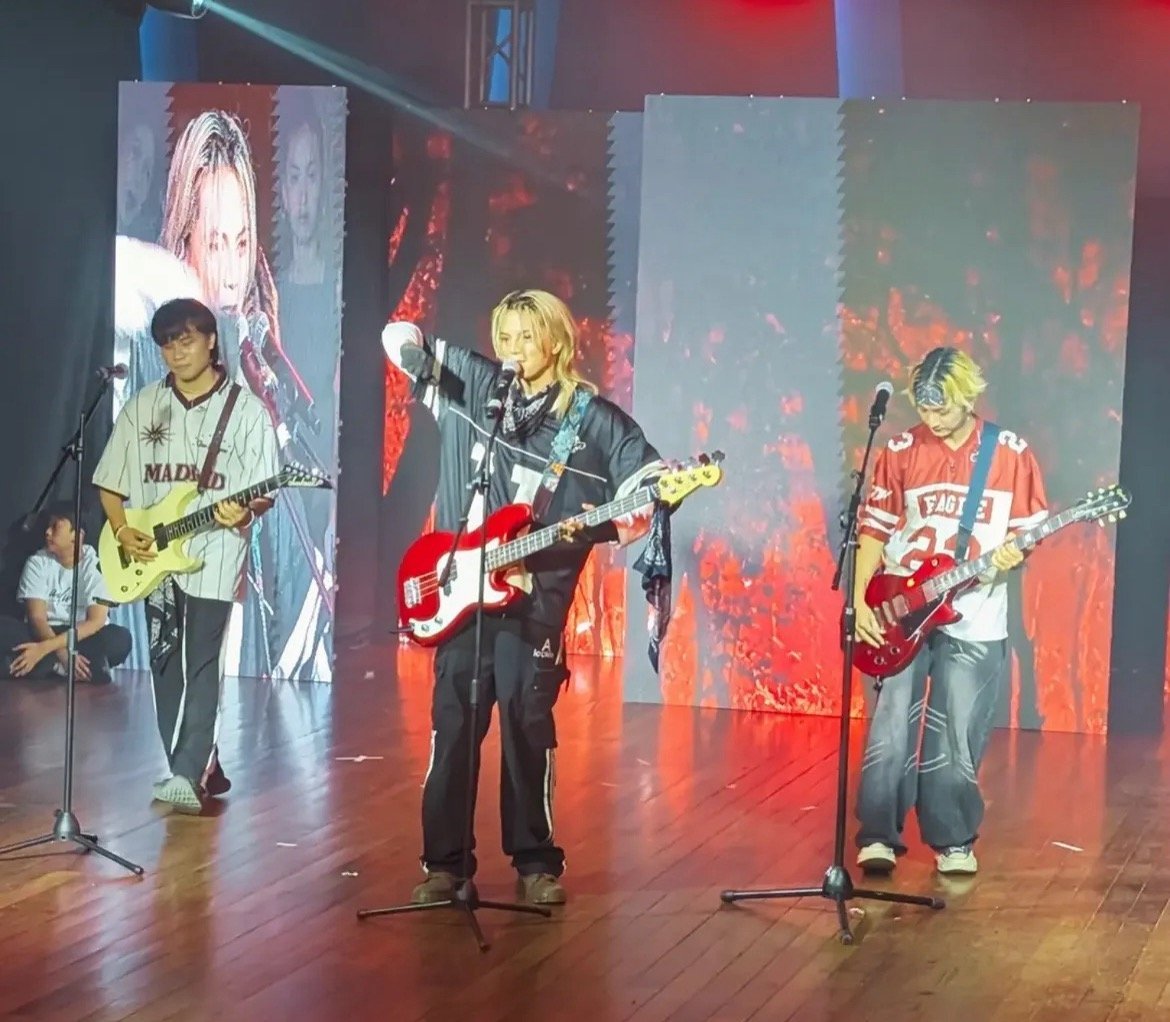 Three musicians performing with guitars on stage under red lighting with large video screens displaying performers in background