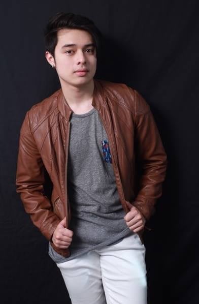 Young man wearing a brown leather jacket over gray t-shirt and white pants, standing against a dark background