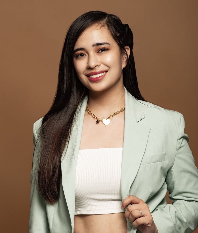 Young woman with long black hair smiling at camera, wearing light green blazer over white crop top and gold chain necklace against beige background