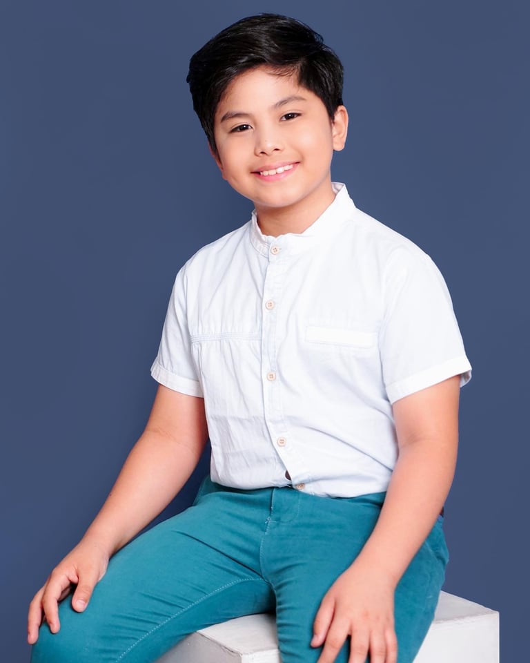 Young boy in white short-sleeved shirt and teal pants sitting and smiling against blue background