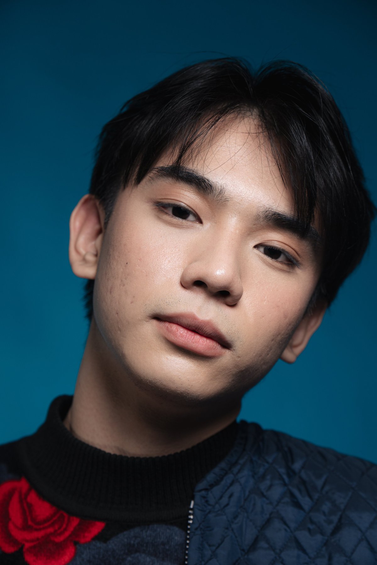Portrait of a young Asian man with black hair wearing a black and red jacket against a teal blue background