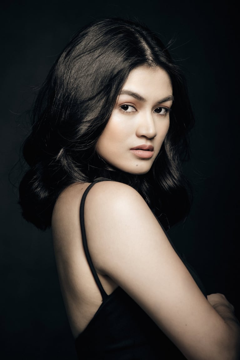 Woman with long dark wavy hair wearing black sleeveless top, looking over shoulder against dark background