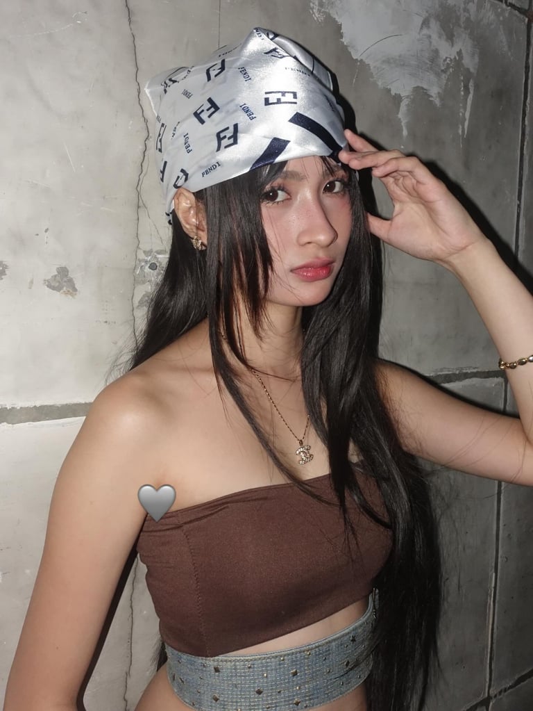 Woman with long black hair wearing a white patterned cap and brown crop top against a weathered gray wall