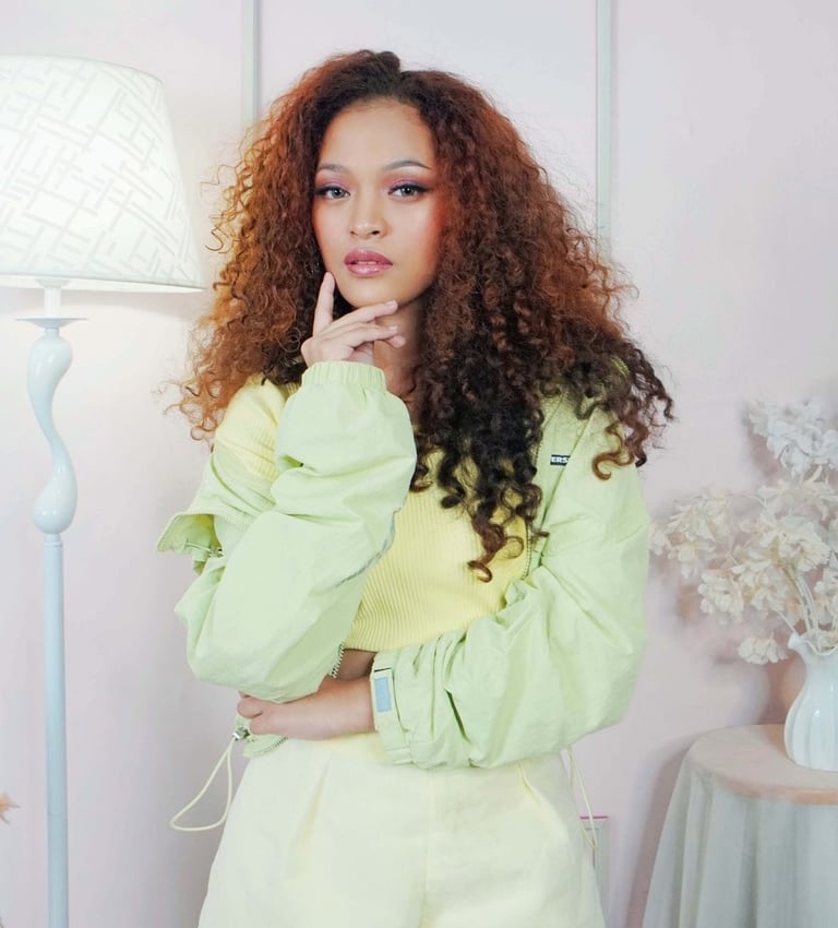 Woman with long curly reddish-brown hair wearing a pale green outfit, posing thoughtfully in a bright room with a lamp and flowers.