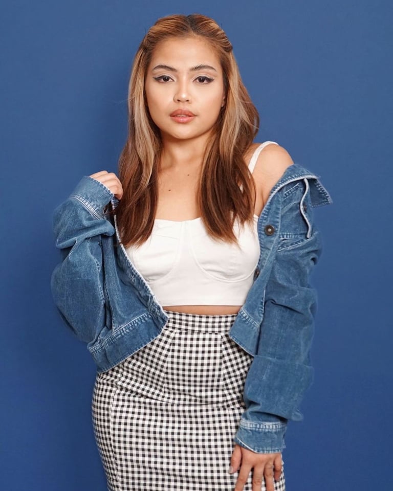 Young woman wearing a white crop top, blue denim jacket, and black and white gingham skirt against a blue background