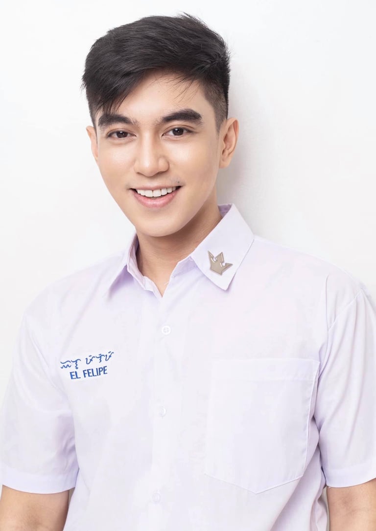 Young man with short dark hair smiling, wearing a light purple shirt with a name tag and collar pin against a white background
