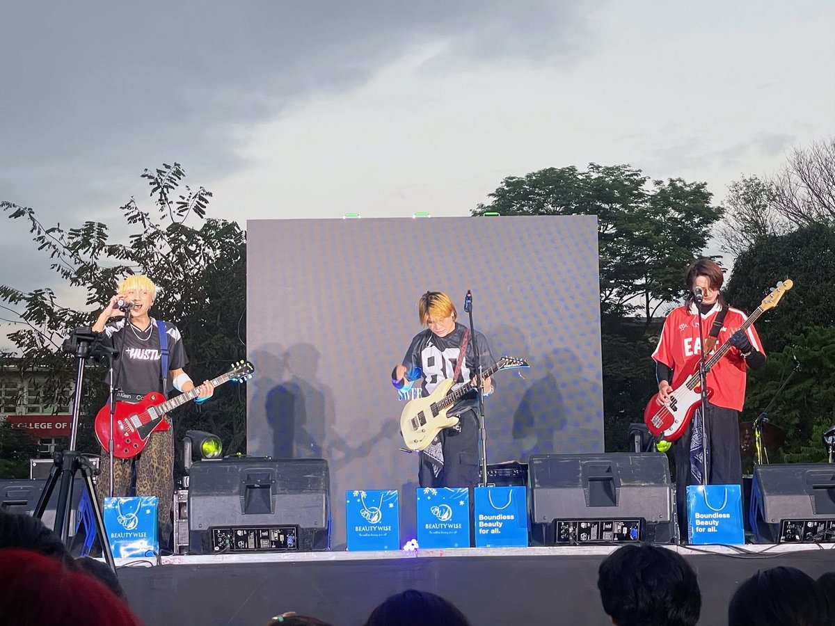Four musicians performing on an outdoor stage with guitars and microphones, large video screen behind them, blue speaker boxes visible.