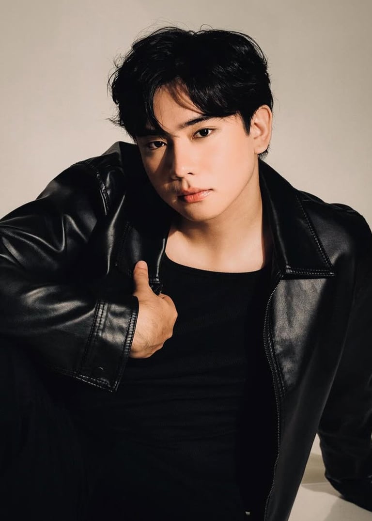 Young man with black hair wearing a black leather jacket over a black t-shirt against a neutral background