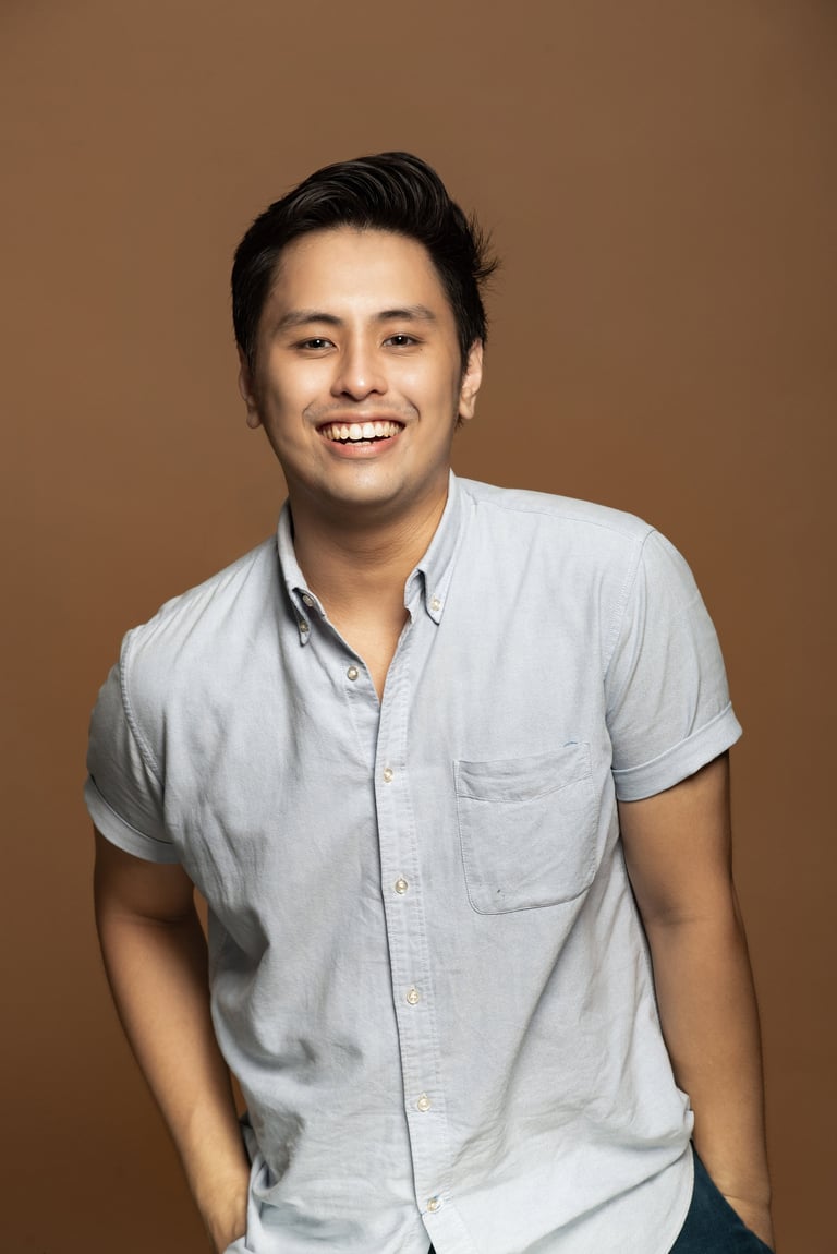 Smiling Asian man wearing a light blue short-sleeved button-up shirt against a tan background