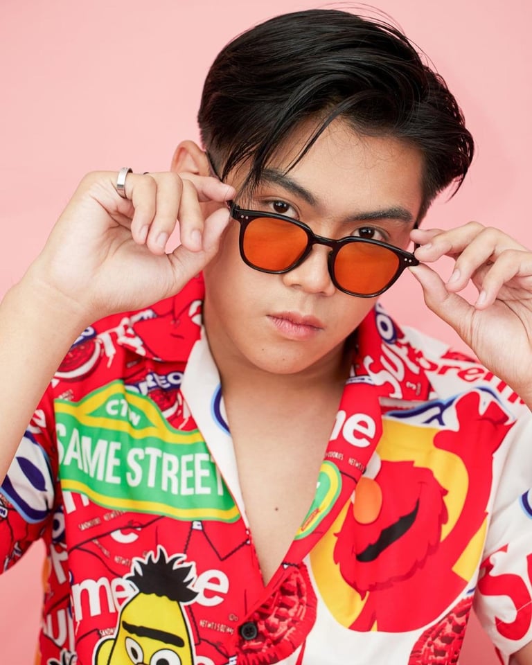 Person wearing orange-tinted sunglasses and a colorful Sesame Street graphic jacket against a pink background