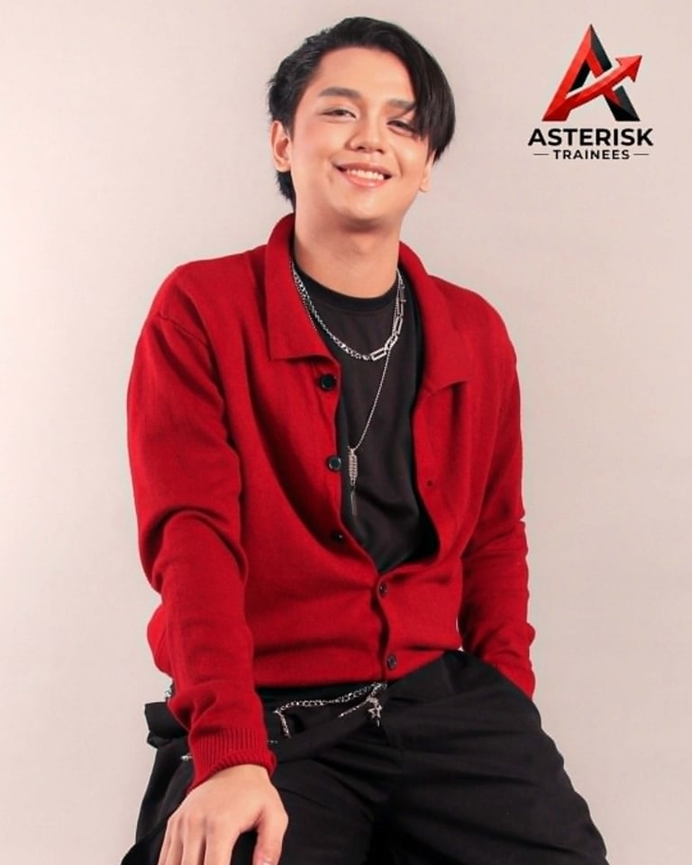 Smiling young person wearing a red cardigan over a black shirt with chains, with the Asterisk Trainees logo in the background