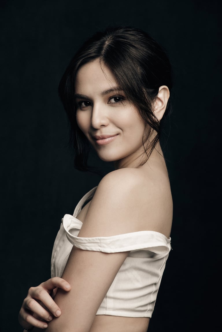 Woman with dark hair wearing off-shoulder white top, looking over shoulder against black background