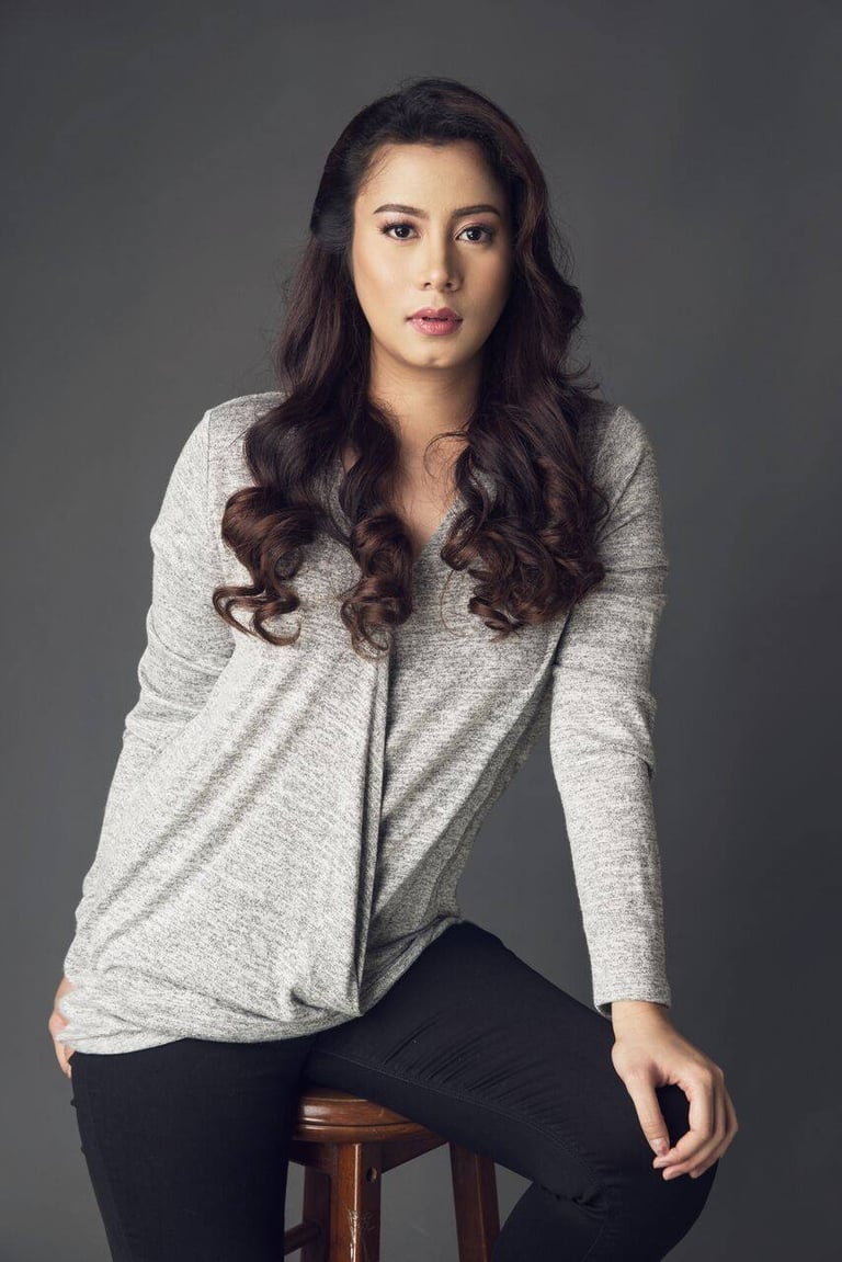 Woman with long dark wavy hair wearing a light gray cardigan and black pants, sitting on a wooden stool against a gray background